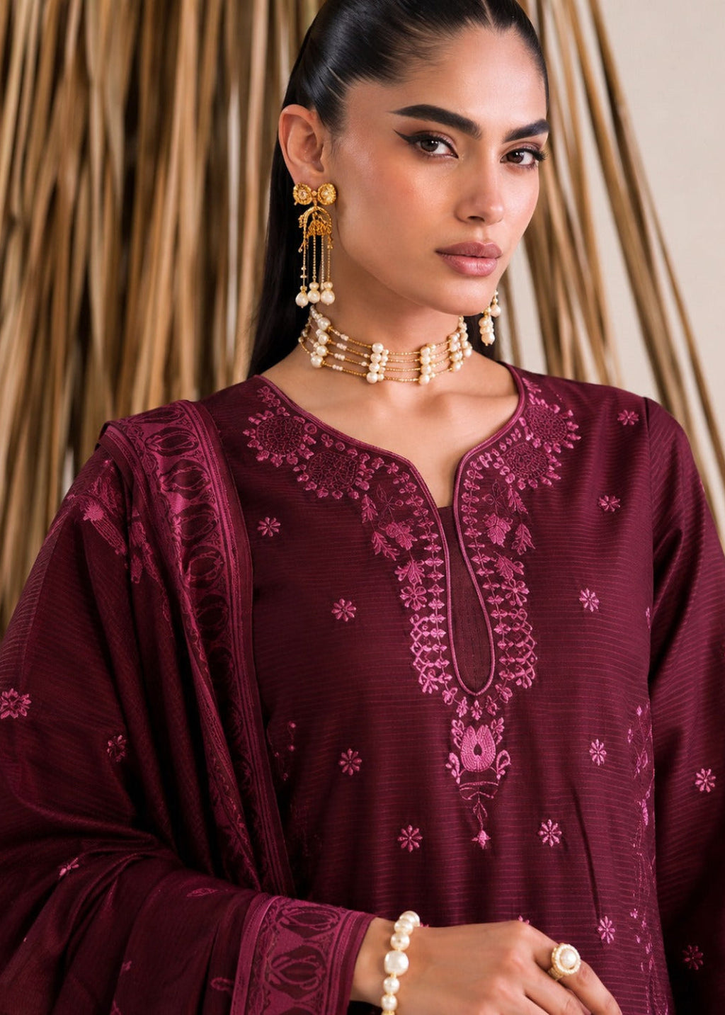 A woman wearing a deep maroon embroidered kurta with a matching dupatta, styled with gold jewelry against a neutral background with vertical wood slats.