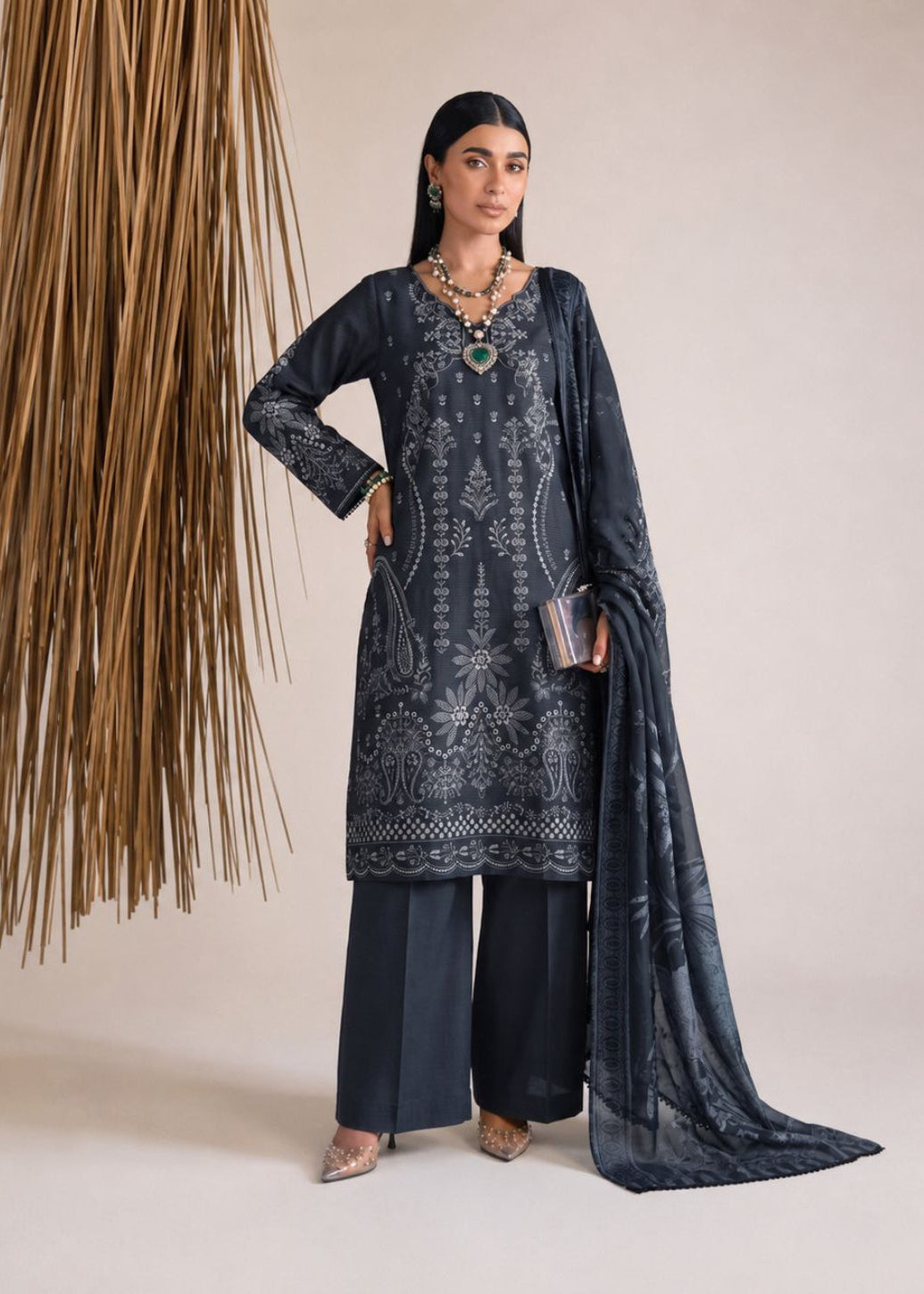A woman models a dark navy blue ethnic salwar kameez suit with intricate white embroidery, matching wide-leg trousers, and a sheer embroidered dupatta, standing against a neutral beige background with a decorative hanging reed arrangement.
