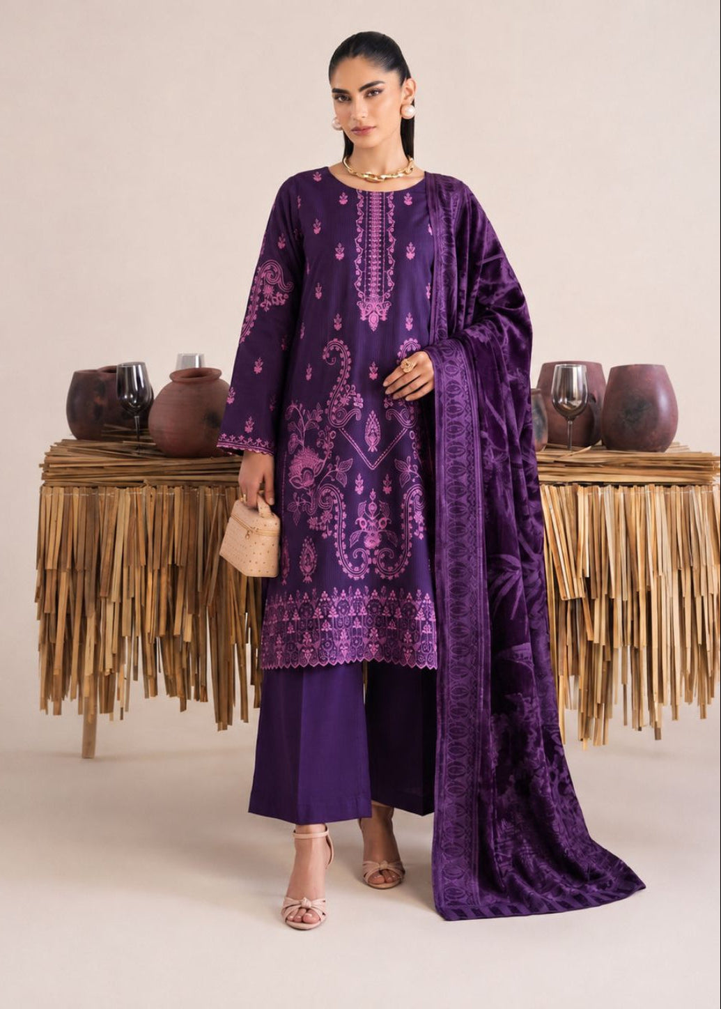 A model stands in a studio wearing a deep purple traditional outfit with matching embroidered kurta, wide-leg trousers, and a draped dupatta. The outfit features intricate lighter purple embroidery and motifs.