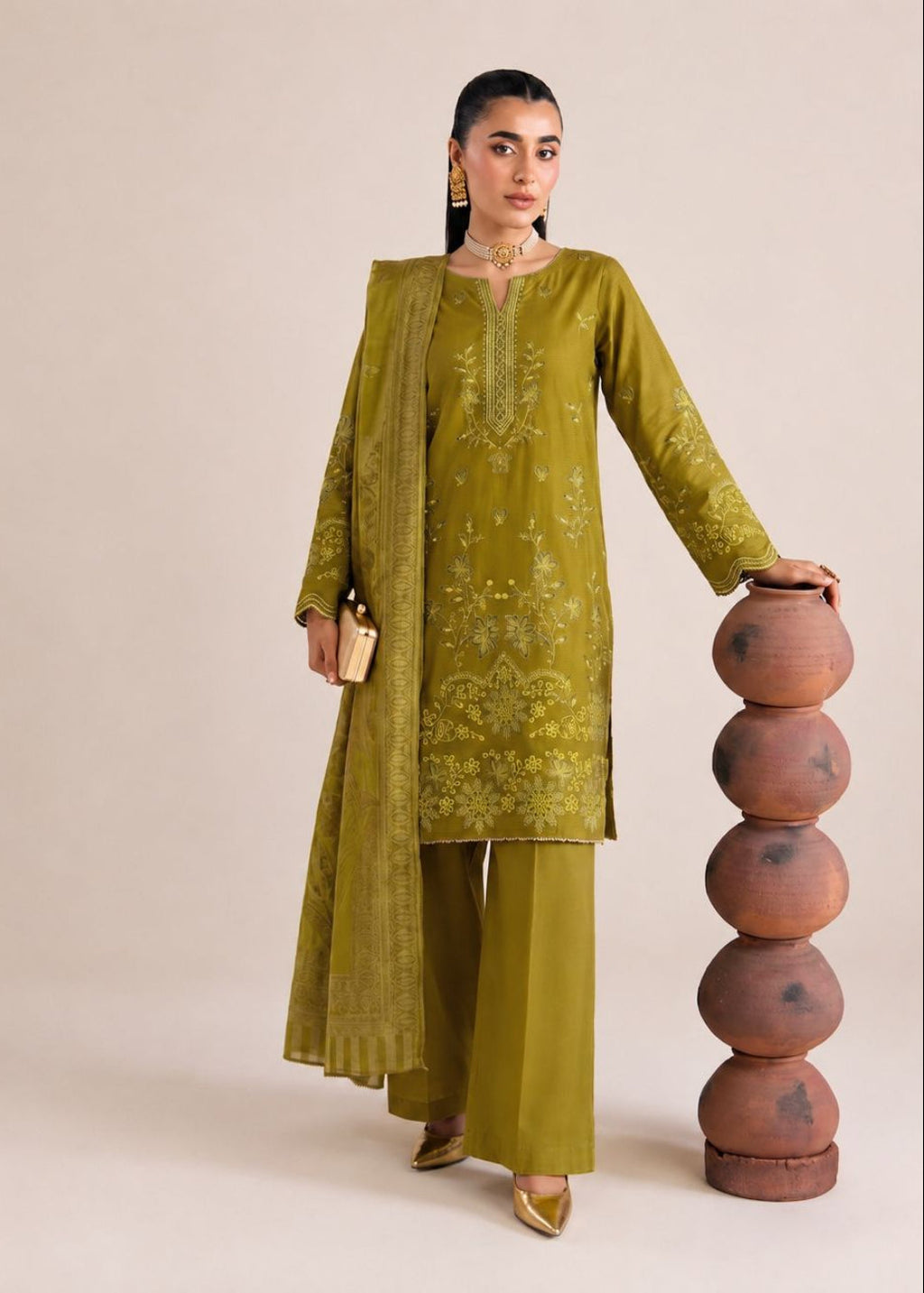 A woman models a three-piece olive green ethnic suit with matching dupatta, featuring gold embroidery on the kurta and trousers, standing next to a stack of terracotta pots against a beige studio background.