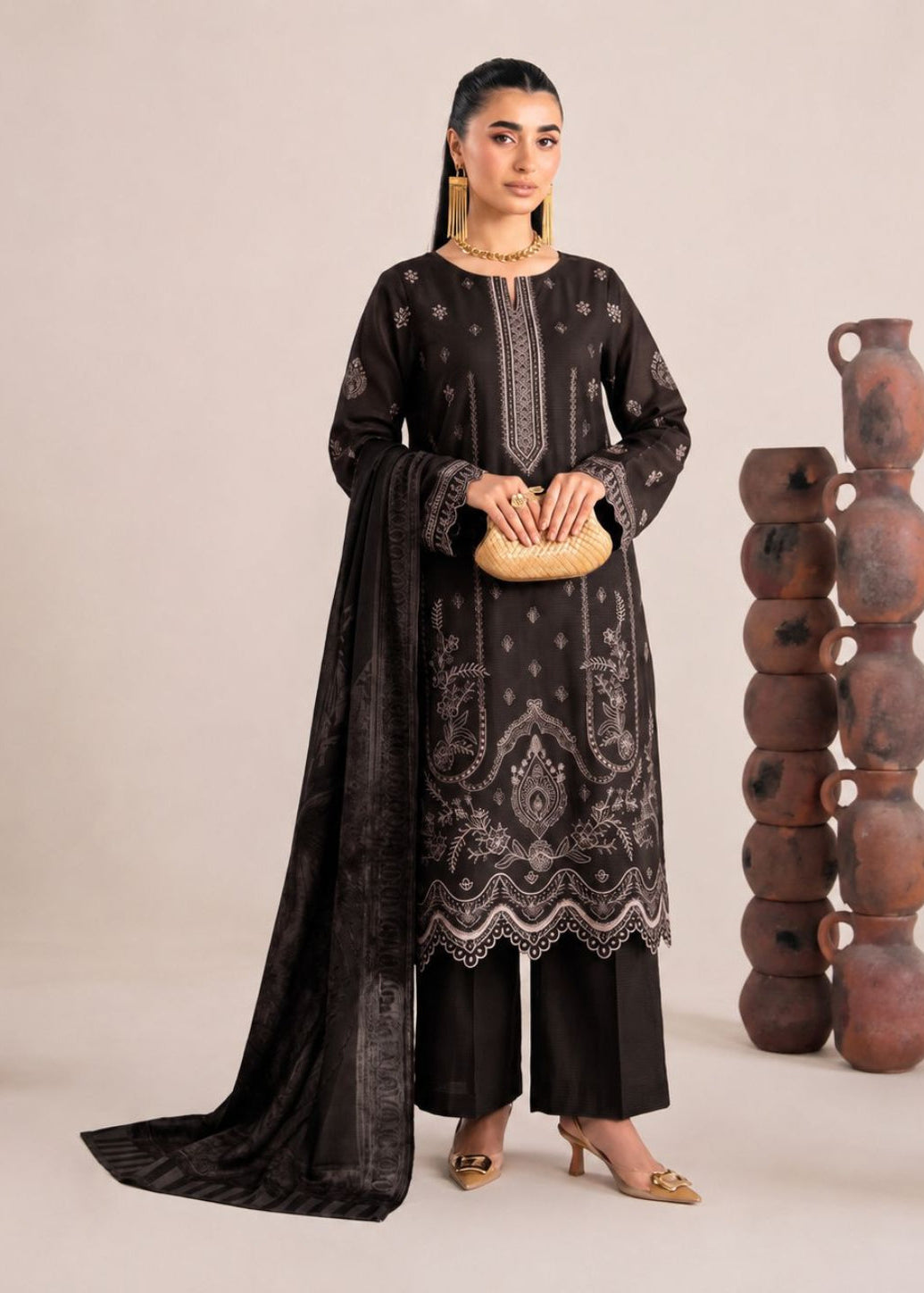 A woman models a black three-piece traditional outfit with intricate silver embroidery, featuring a long kurta, matching trousers, and a draped dupatta. She is standing in a studio setting with decorative stacked terracotta pots on either side.