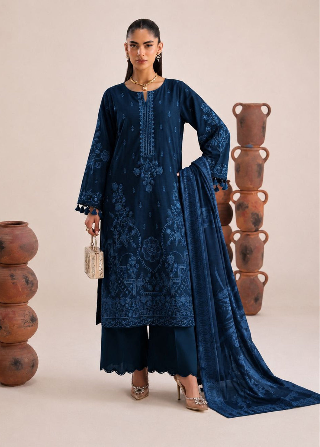 A woman models a traditional South Asian outfit consisting of a navy blue embroidered kurta, matching wide-leg pants, and a coordinating dupatta. She is standing in a studio with decorative stacked terracotta pots on either side, against a solid beige background.