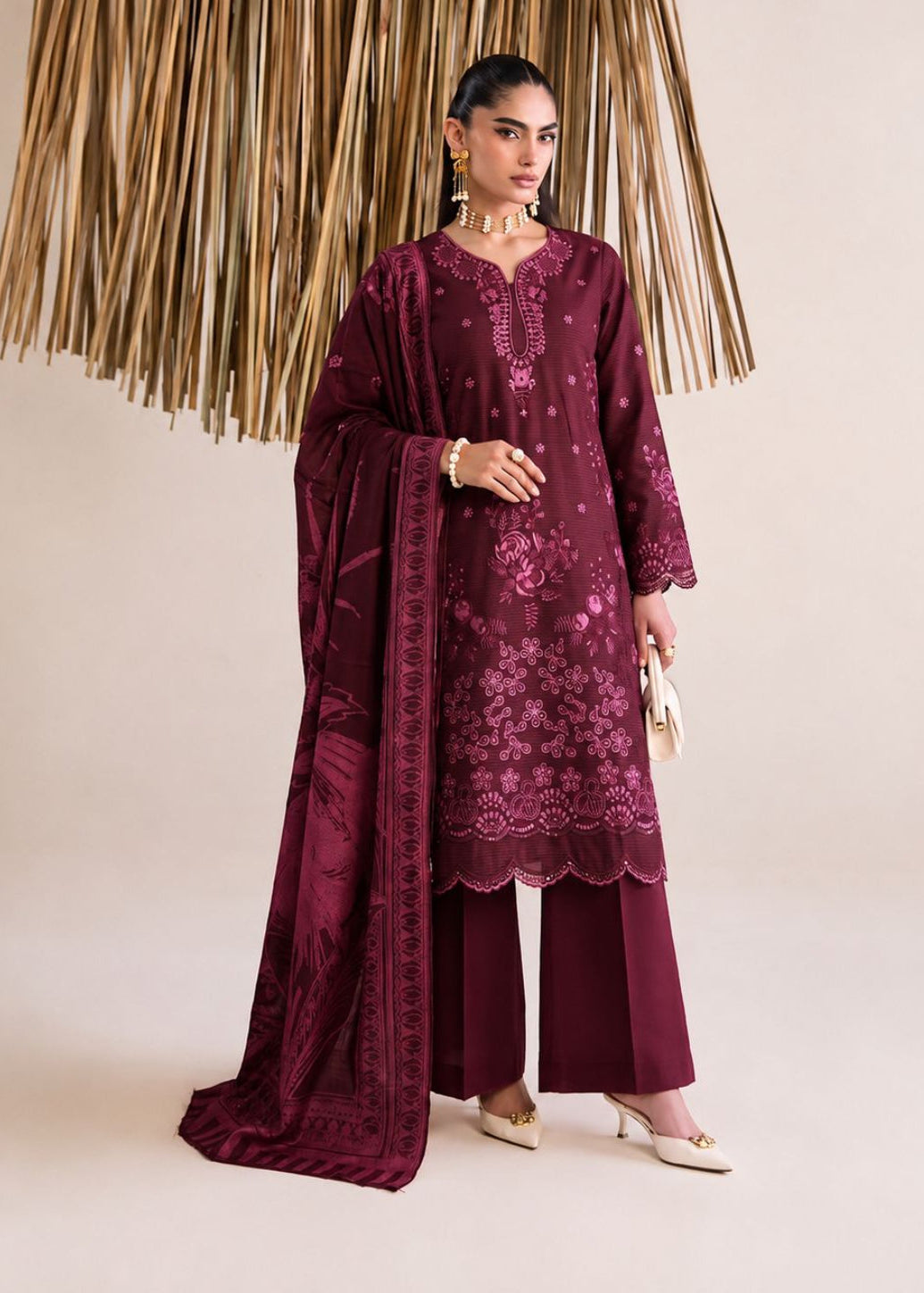 A woman models a three-piece traditional outfit in deep maroon with floral embroidery, featuring a long kurta, wide-leg pants, and a matching dupatta, set against a neutral studio backdrop with a decorative gold hanging.