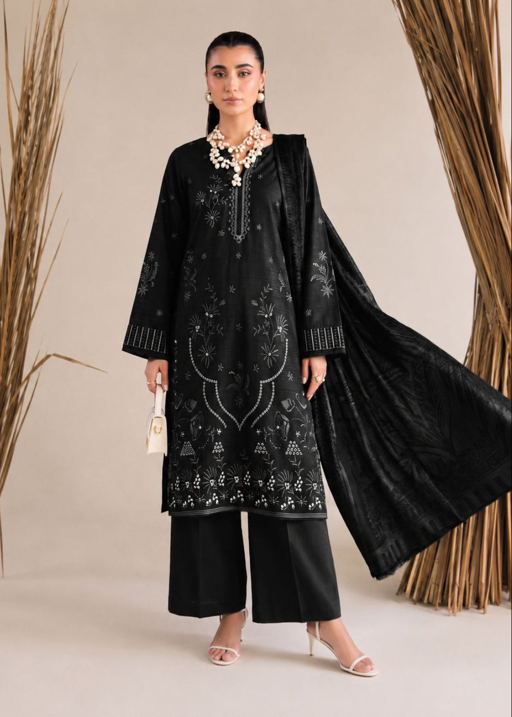 A woman models a black traditional South Asian outfit with intricate silver embroidery, wide-leg pants, a matching black dupatta, and a statement necklace, set against a neutral beige background with dried palm branches.