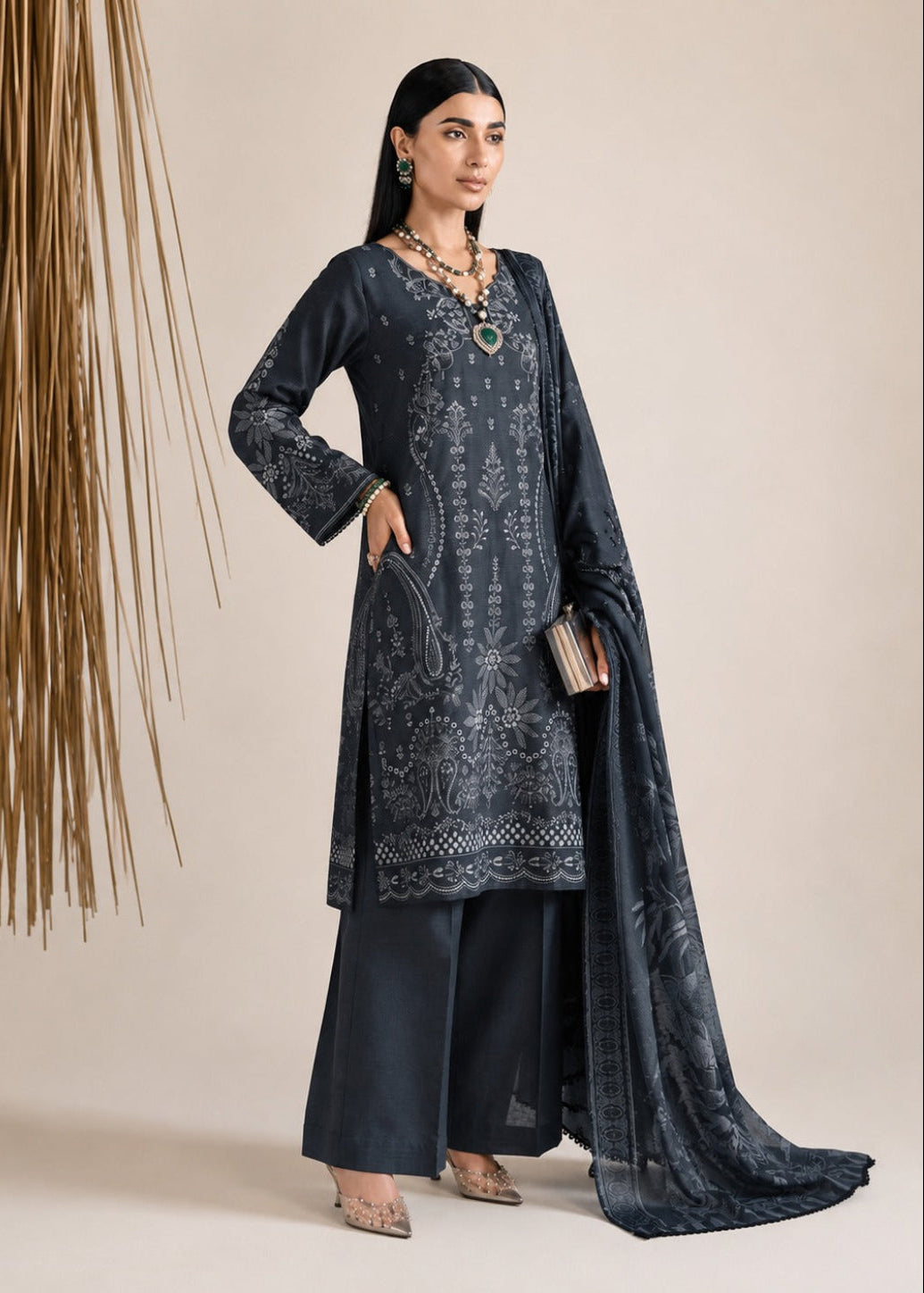 A woman models a dark navy blue ethnic salwar kameez suit with intricate silver embroidery, matching wide-leg trousers, and a sheer embroidered dupatta, set against a neutral beige background with a decorative dried palm frond element on the left.