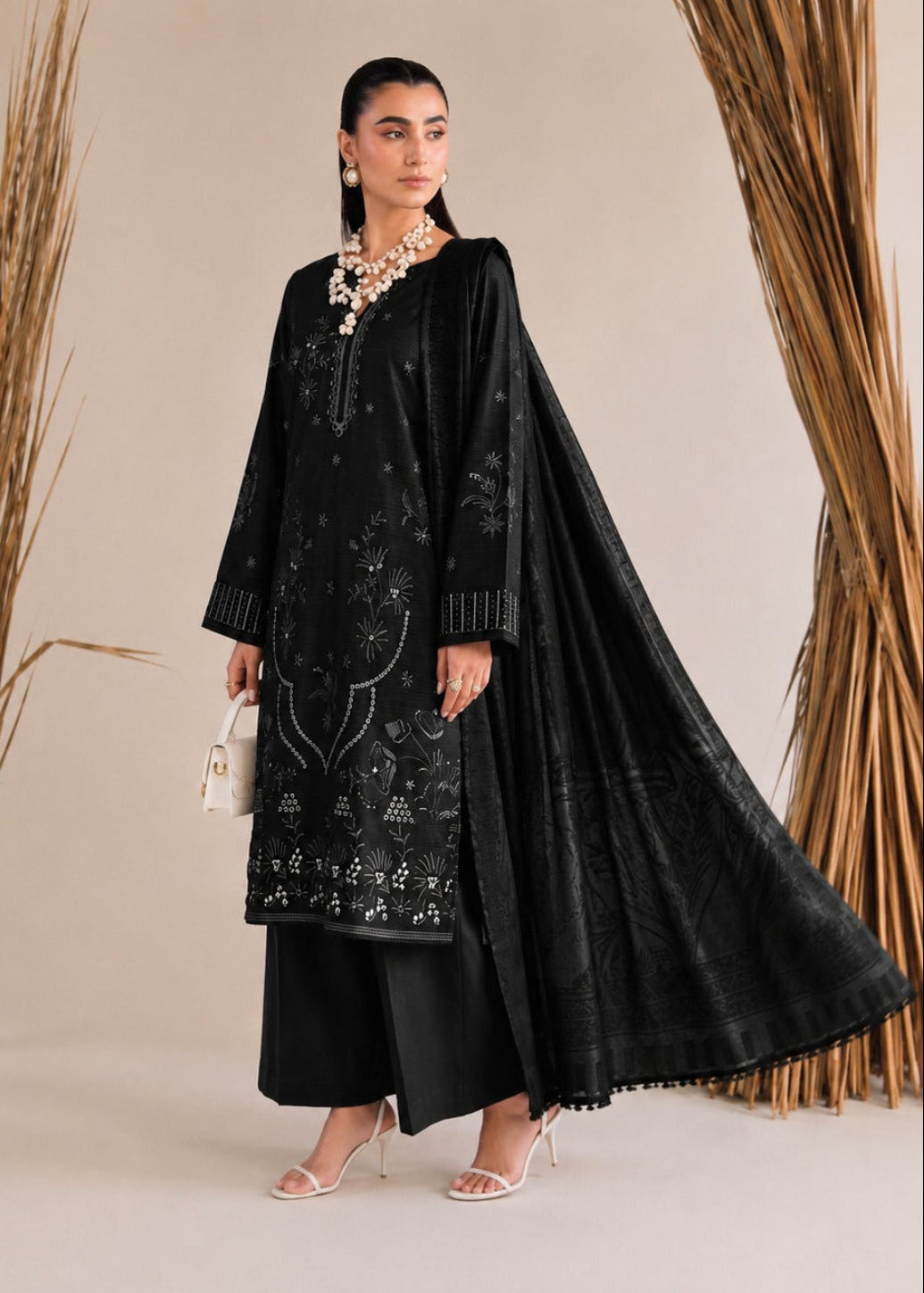 A woman models a black embroidered traditional outfit with a matching sheer dupatta, black pants, and white heels, standing against a beige backdrop with dried decorative grasses.
