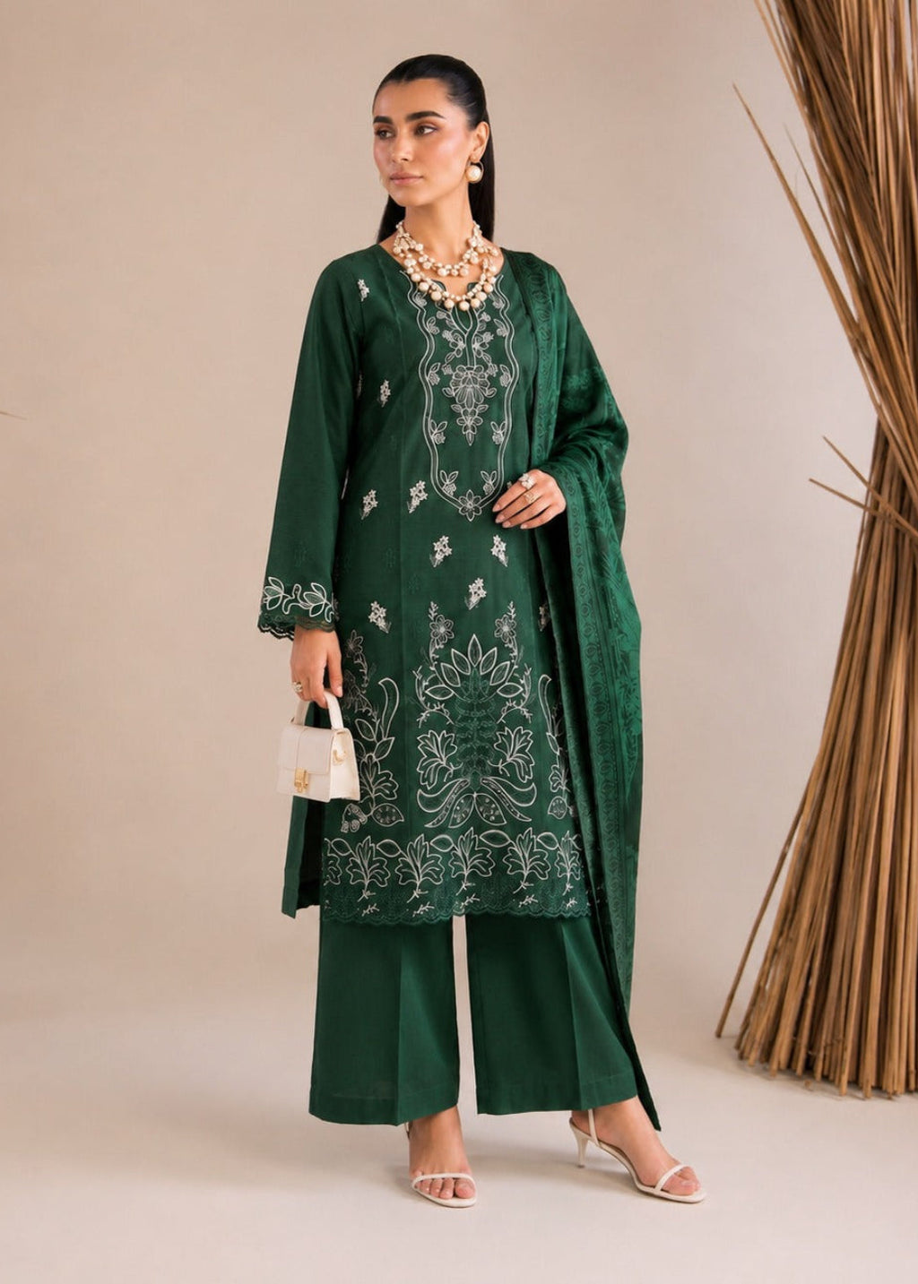 A woman models a dark green traditional outfit with white embroidery, matching wide-leg pants, a draped jacket, and a chunky gold necklace, standing against a beige studio backdrop with decorative dried palm leaves.