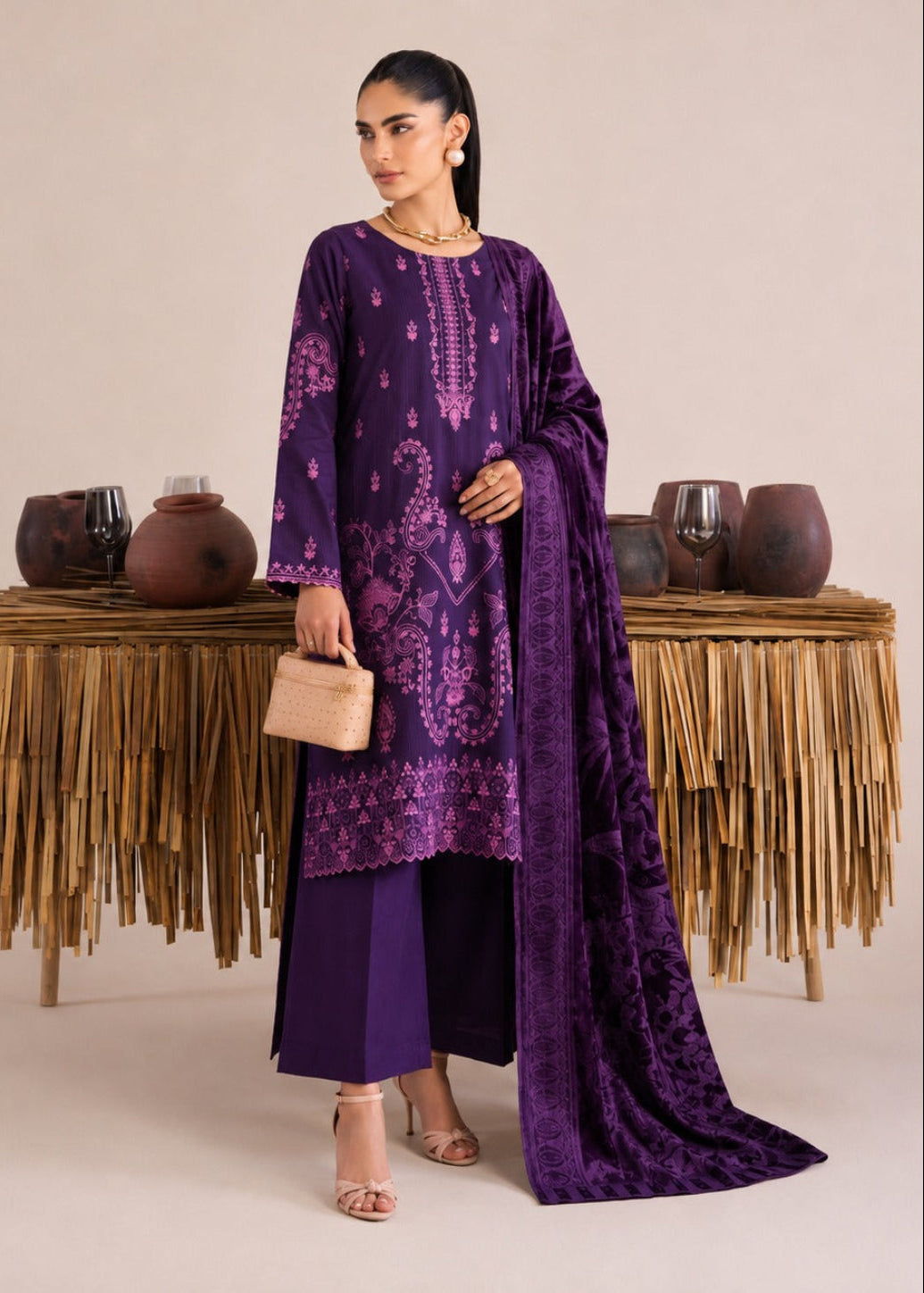 A woman models a deep purple traditional South Asian outfit with matching pants and a long dupatta, standing in front of a beige studio backdrop with decorative woven tables and ceramic pots.