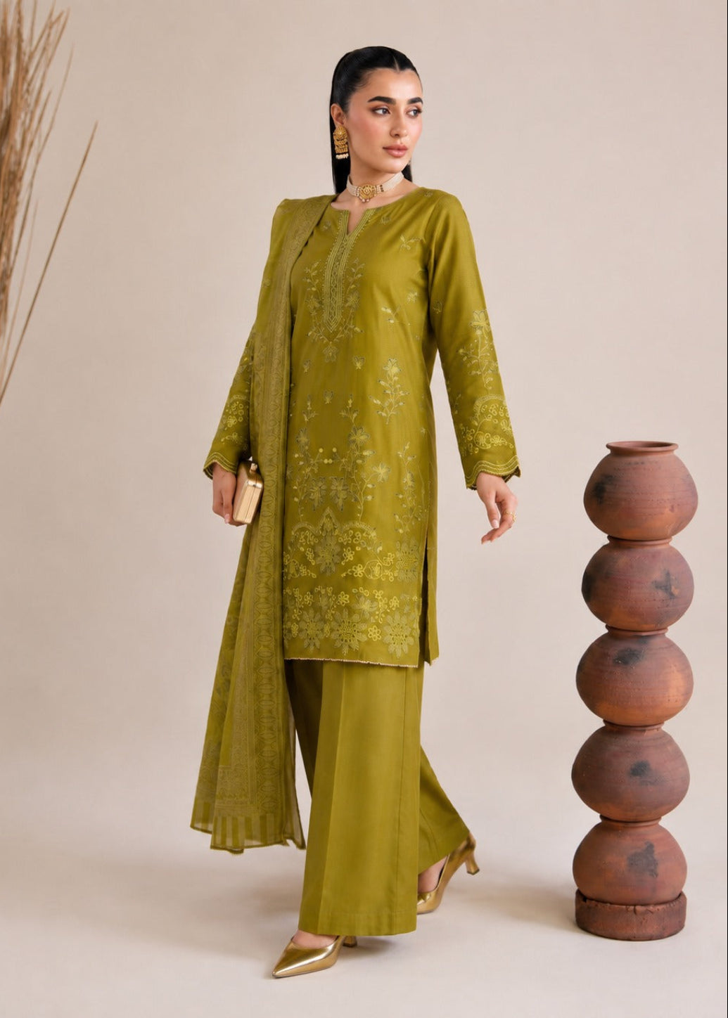 A woman models an olive green traditional South Asian outfit consisting of a long-sleeved embroidered kurta, matching wide-leg trousers, and a sheer dupatta, set against a neutral beige background with decorative props.