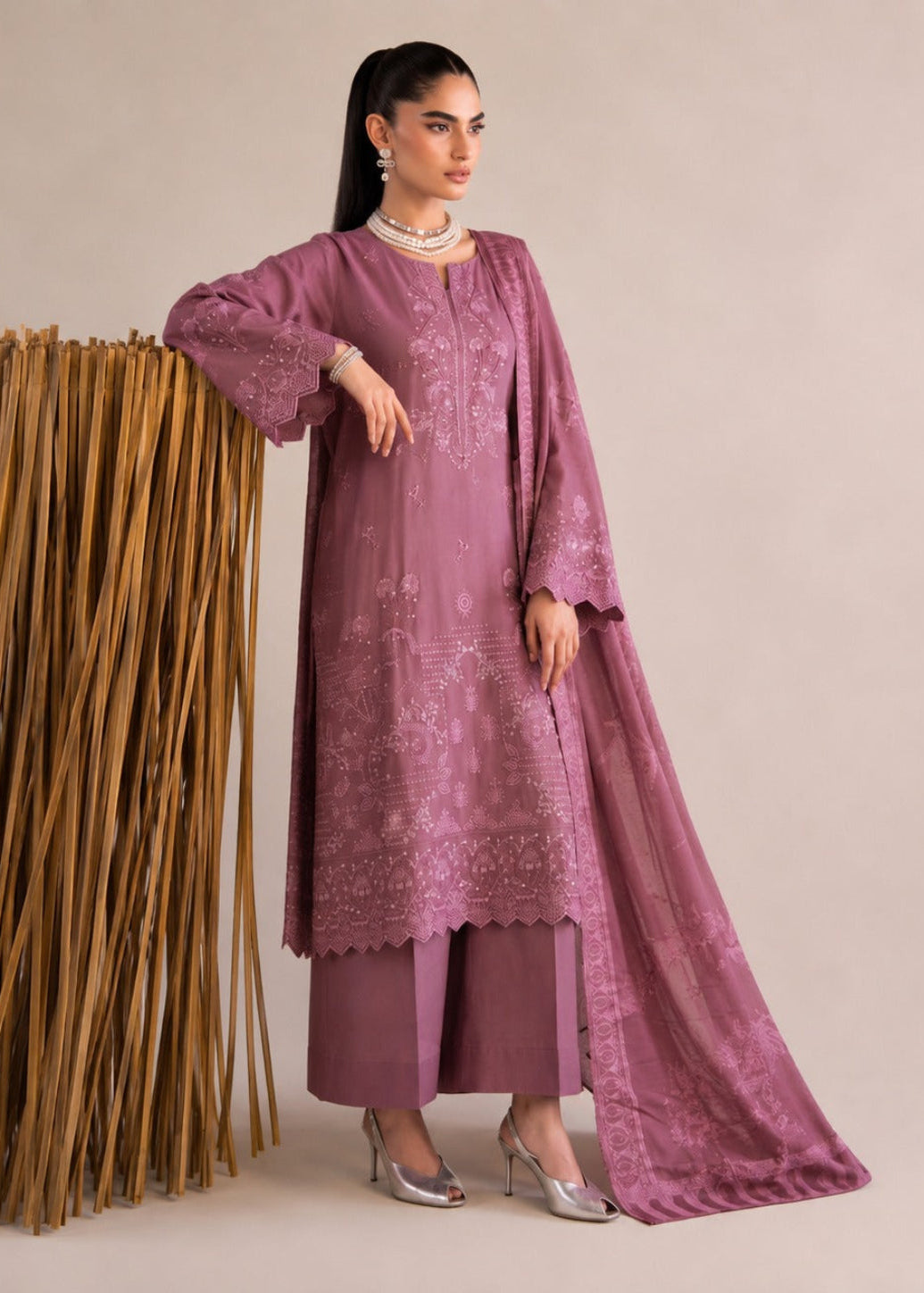 A woman models a three-piece traditional outfit in dusty mauve, featuring a long embroidered kurta, matching wide-leg trousers, and a sheer dupatta. She stands next to a decorative arrangement of dried palm leaves against a solid beige studio background.