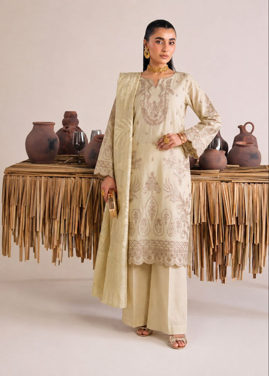 A woman models a traditional beige embroidered salwar kameez suit with a matching dupatta, standing in front of a styled studio backdrop with decorative clay pots and a fringed table runner.
