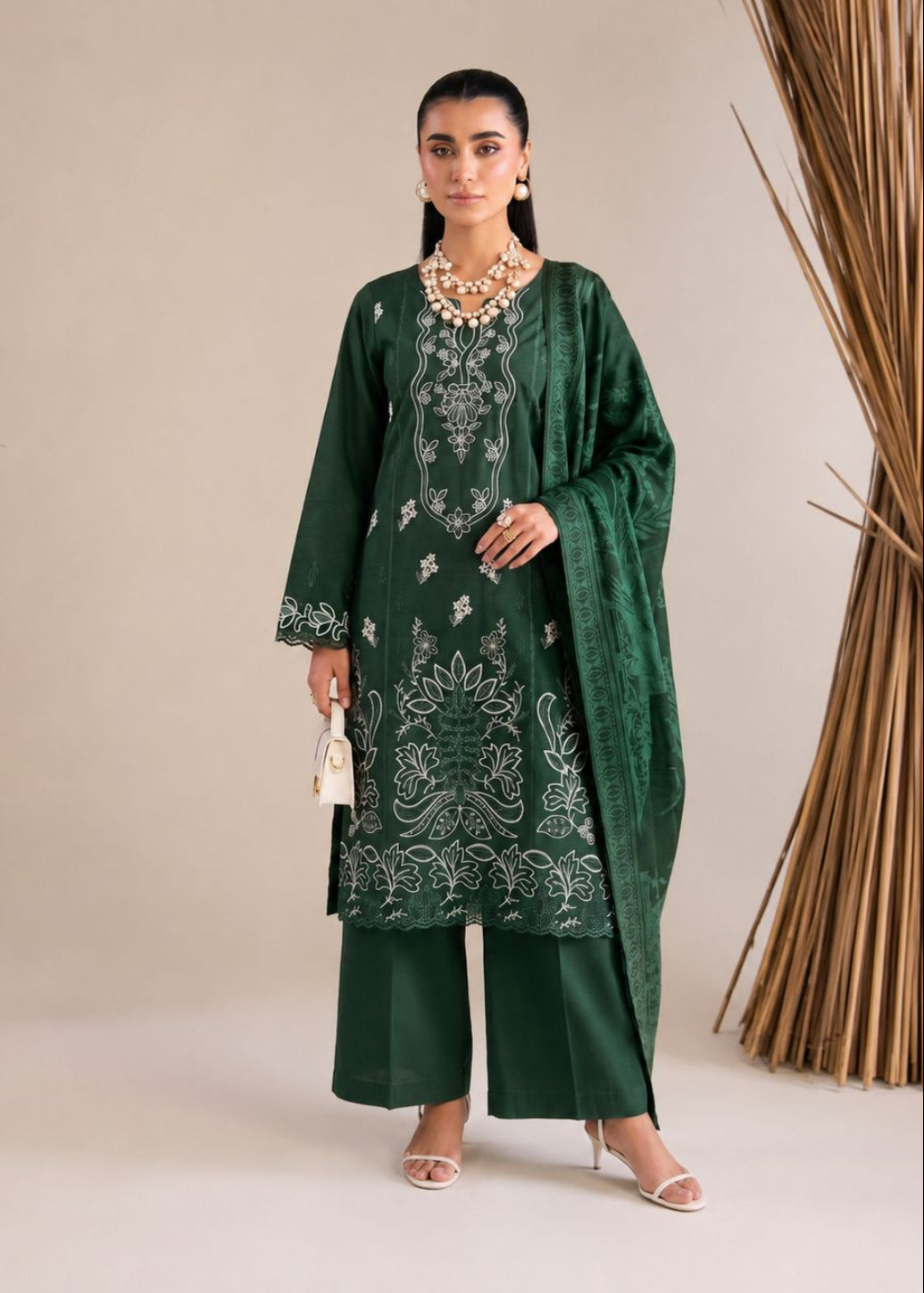A woman models a dark green traditional South Asian outfit with white embroidery, matching wide-leg trousers, and a coordinating dupatta, set against a neutral beige background with decorative dried palm leaves.