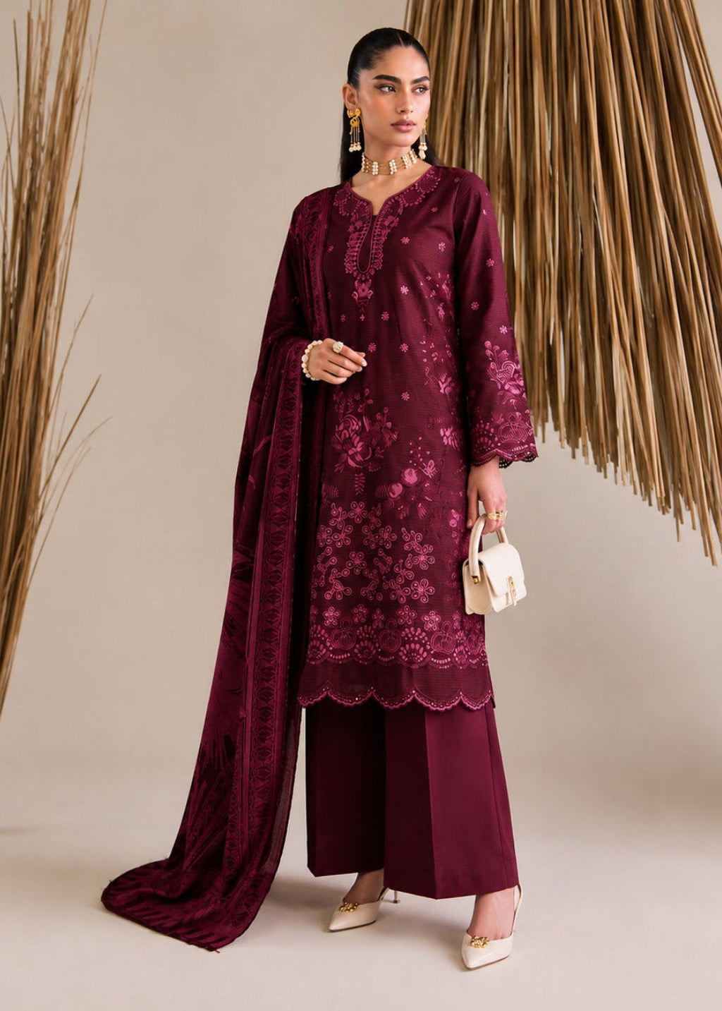 A woman models a deep maroon three-piece ethnic suit with matching dupatta, featuring floral embroidery, lace borders, and wide-leg trousers, styled with gold jewelry and a small white clutch against a beige backdrop with dried palm leaves.
