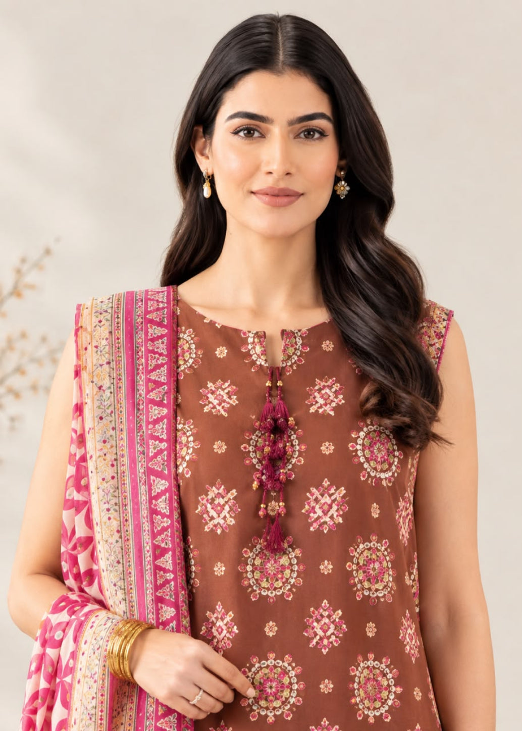 A woman modeling a brown 3-piece salwar kameez suit with pink and gold floral embroidery, paired with a pink patterned dupatta.