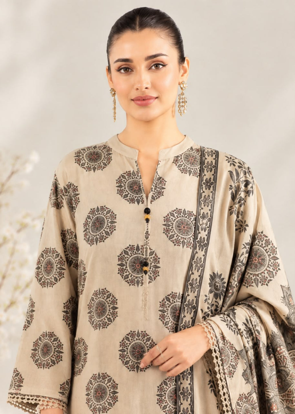 A woman modeling a beige three-piece traditional outfit with dark brown circular floral prints and a matching printed dupatta, set against a soft neutral background.