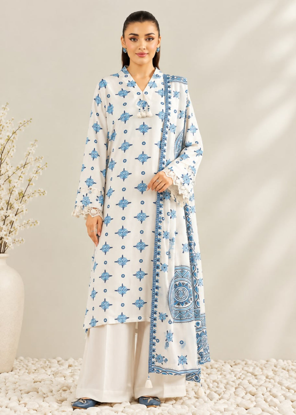 A woman models a traditional white three-piece suit set featuring light blue printed patterns, including a long kurta, wide-leg trousers, and a matching dupatta, set against a neutral beige background with decorative dried flowers.