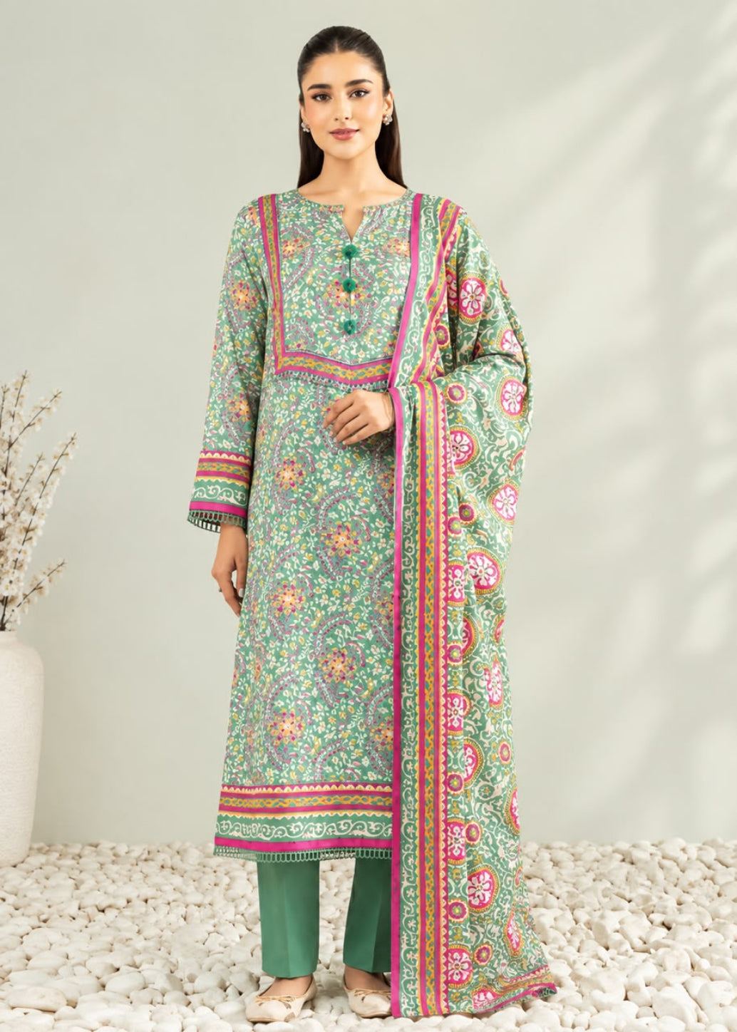 A woman models a traditional three-piece salwar kameez suit set. The set features a mint green printed kurta with pink and orange floral motifs, matching green trousers, and a coordinating dupatta with a vibrant pink and green border.