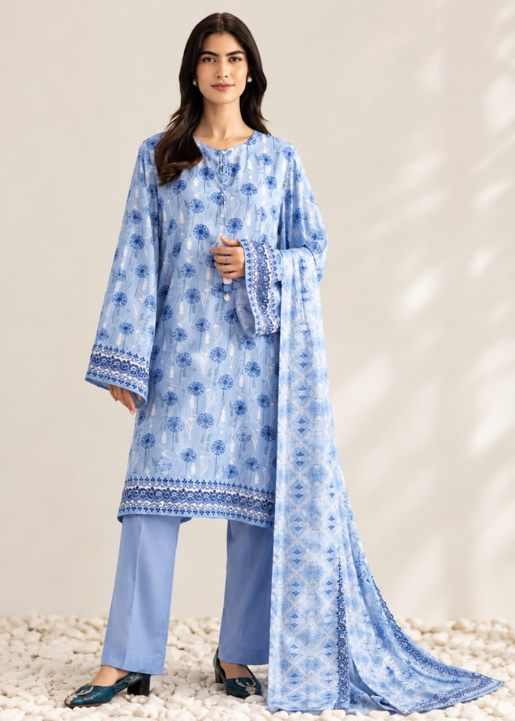 A woman models a light blue three-piece traditional outfit with a matching dupatta. The set features a long-sleeved tunic with blue floral prints and embroidered borders, light blue pants, and a matching sheer dupatta with a blue geometric pattern.