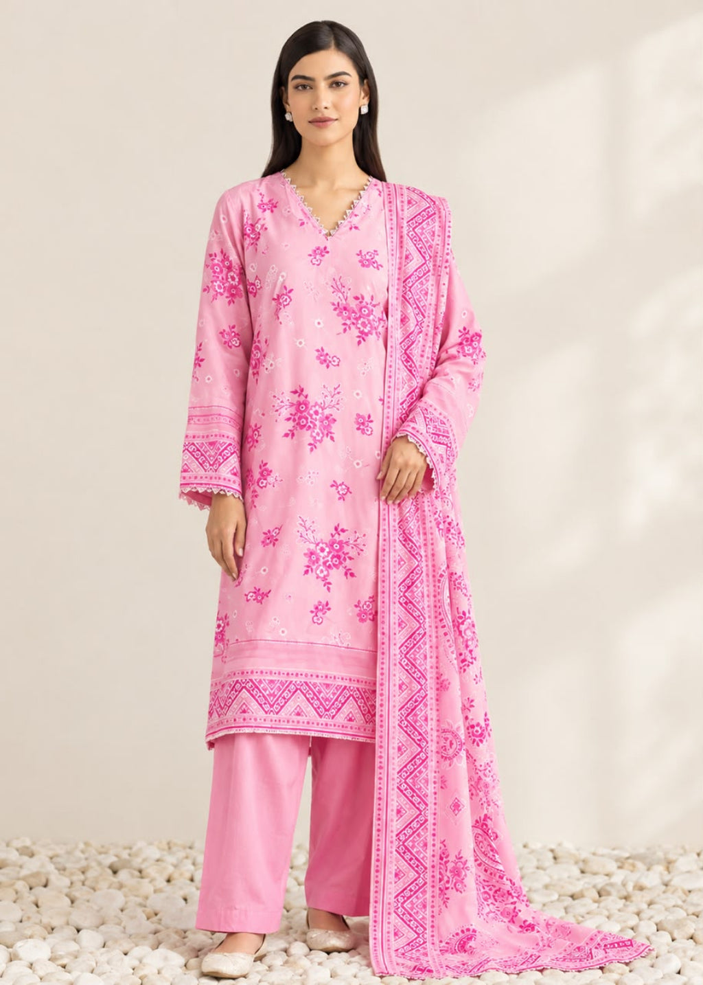 A woman modeling a pink traditional outfit with matching pants and a draped dupatta, featuring bright pink floral embroidery and geometric border patterns, set against a neutral beige background with white pebbles.