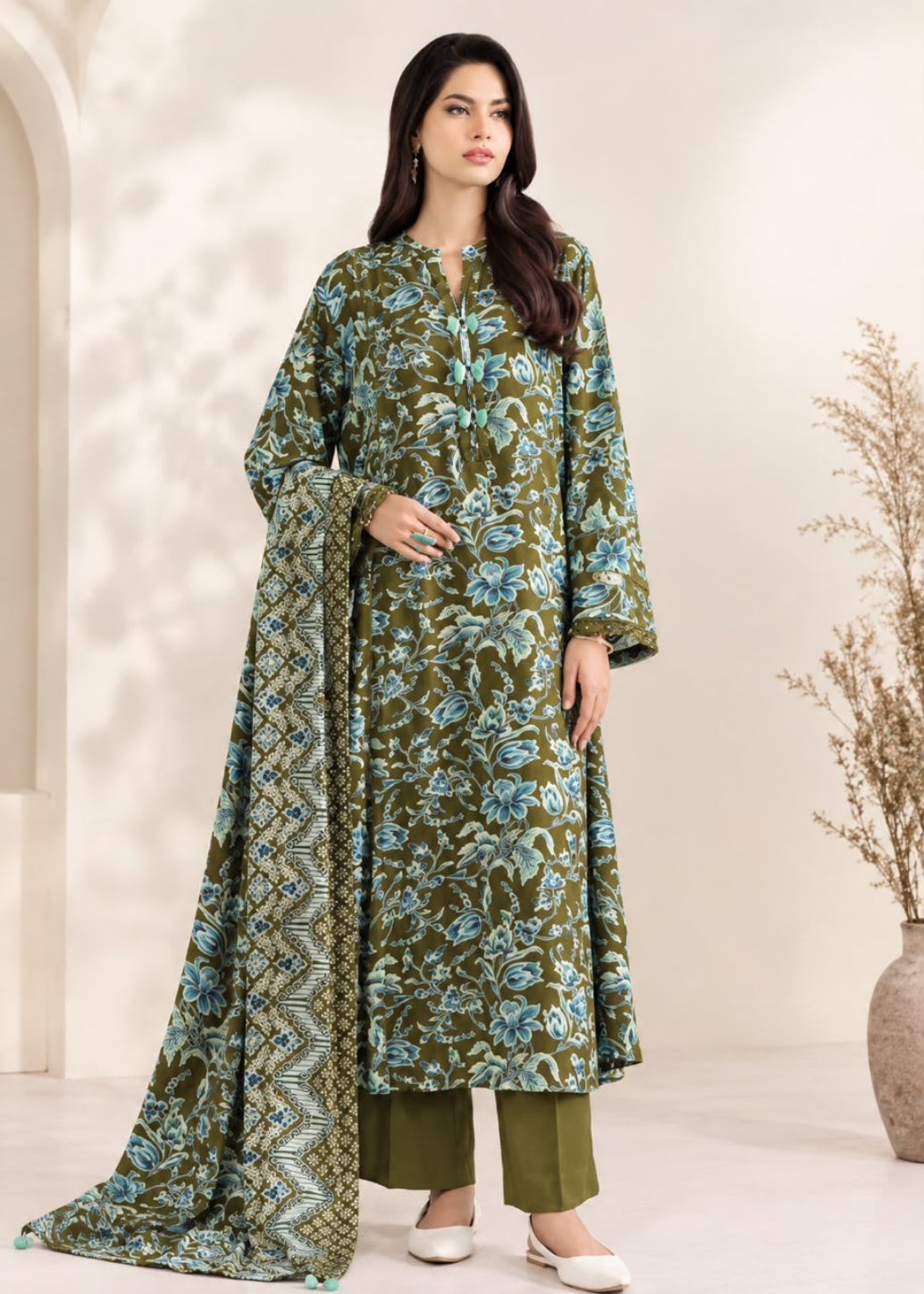 A woman models a three-piece olive green salwar kameez suit with a blue and white floral print, featuring a long kurta, matching trousers, and a coordinating dupatta.