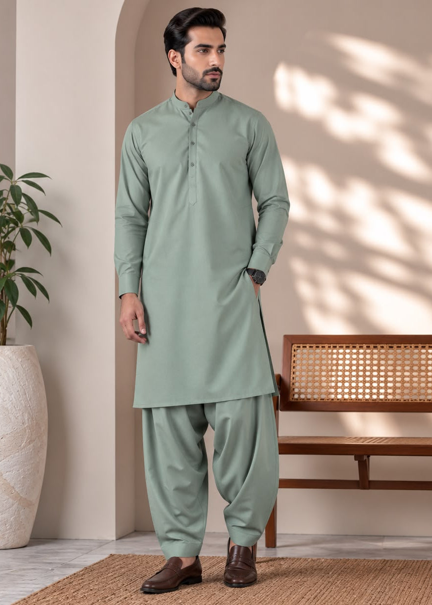 A man wearing a sage green traditional Indian kurta and pant set, standing in a styled indoor setting with a wooden bench, potted plant, and textured rug.