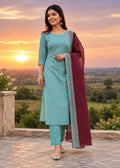 A woman stands outdoors at sunset wearing a light teal three-piece salwar kameez suit with a matching dupatta. The suit features small white floral prints, and the dupatta has a deep maroon border with a silver and black geometric pattern.