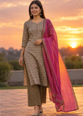 A woman modeling a traditional Indian salwar kameez suit set outdoors at sunset. The set features a beige printed kurta, olive green trousers, and a vibrant magenta dupatta with gold borders.