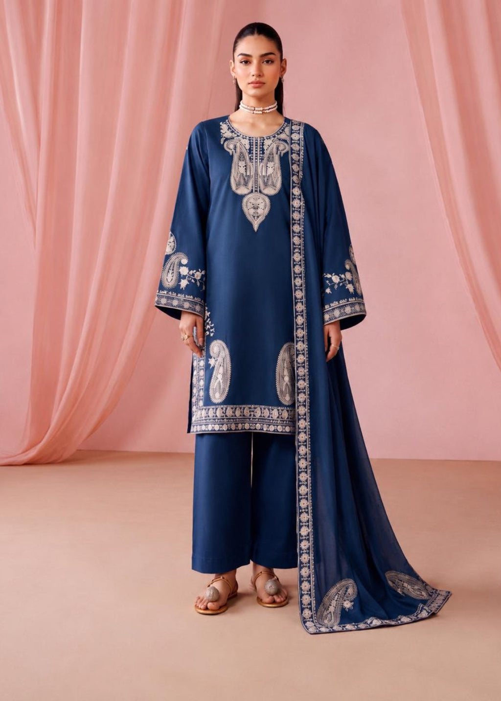 A woman models a navy blue traditional outfit with intricate silver embroidery, matching wide-leg pants, and a long draped scarf, set against a soft pink draped fabric backdrop.