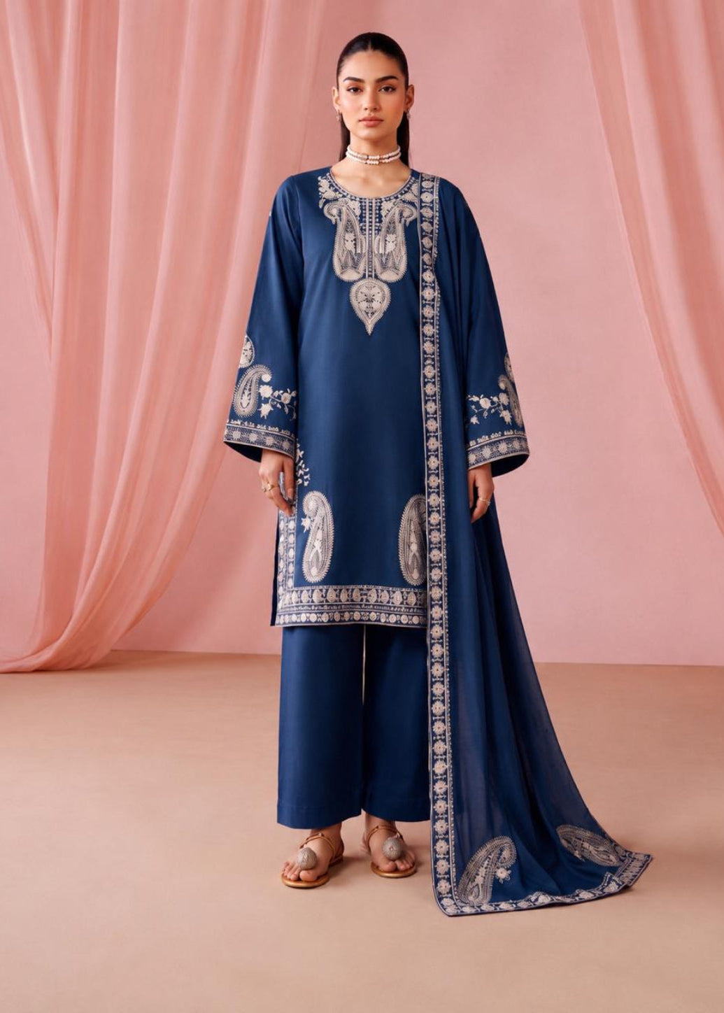 A woman models a navy blue traditional outfit with intricate silver embroidery, matching wide-leg pants, and a long draped scarf, set against a soft pink draped fabric backdrop.