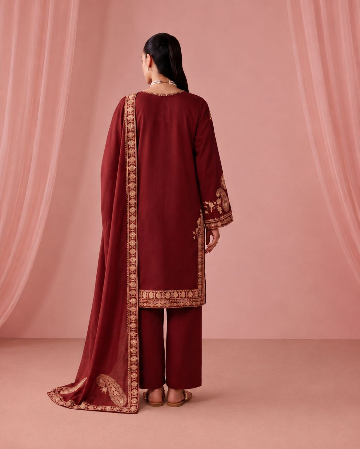 A woman stands with her back to the camera wearing a deep maroon traditional outfit with gold embroidery and a matching dupatta, set against a soft pink draped backdrop.
