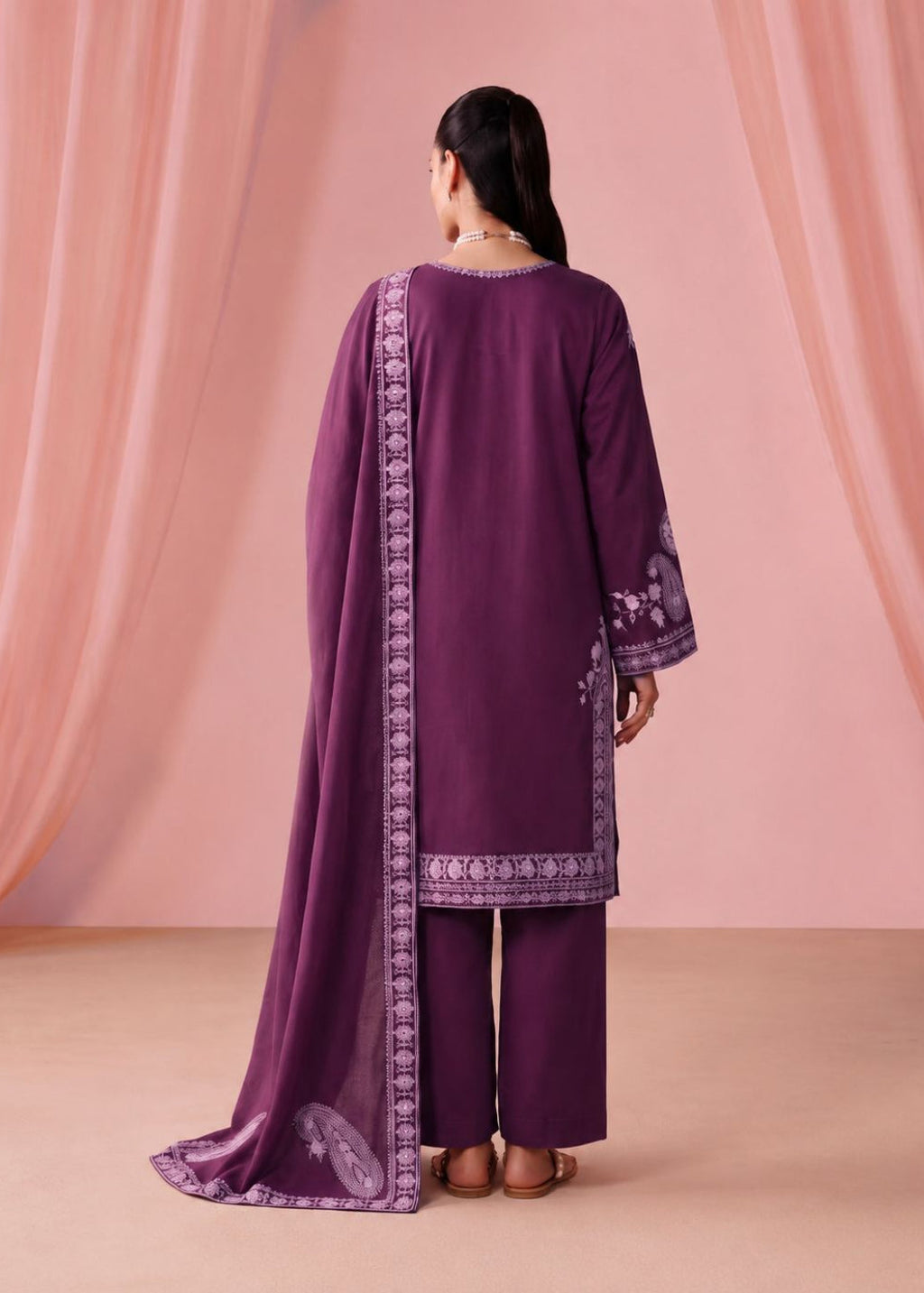 A woman stands with her back to the camera wearing a deep purple traditional outfit with matching wide-leg pants and a draped dupatta featuring light purple border embroidery, set against a soft pink draped backdrop.