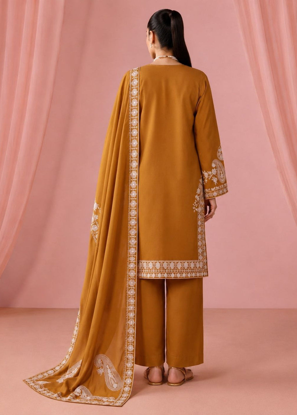 A woman stands with her back to the camera wearing a mustard yellow traditional outfit with white embroidered borders and a matching dupatta, set against a pink draped backdrop.