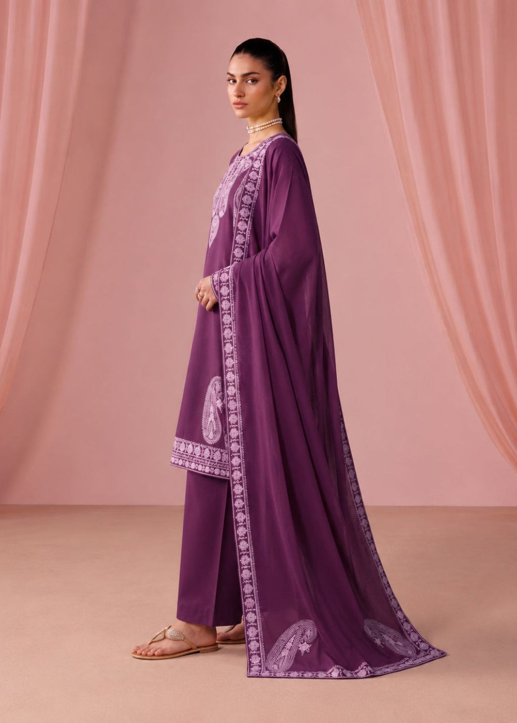 A woman models a traditional purple salwar kameez suit with a matching dupatta. The outfit features light purple embroidery on the neckline, sleeve cuffs, and border, with paisley motifs near the hem.