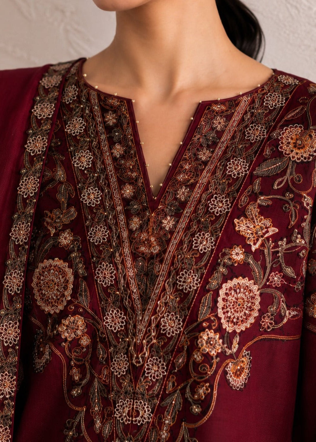 Close-up of a person wearing a deep maroon traditional embroidered outfit with intricate floral embroidery in gold, beige, and brown tones, paired with a matching maroon dupatta against a light textured wall.