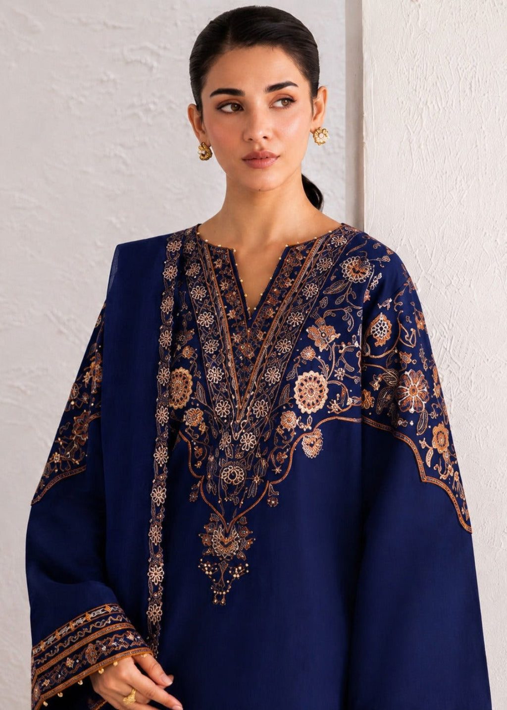A woman modeling a navy blue traditional outfit with intricate gold and beige embroidery, set against a textured off-white wall.