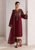 A woman models a deep maroon three-piece salwar kameez suit with intricate gold and floral embroidery on the neckline, sleeves, and border, paired with cream pants and brown sandals, standing against a textured white wall.
