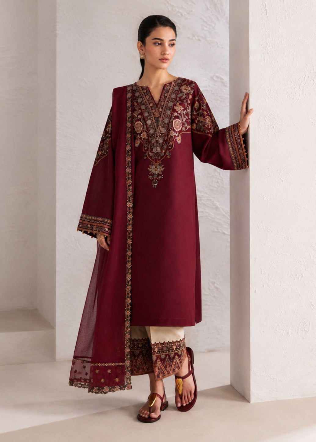 A woman models a deep maroon three-piece salwar kameez suit with intricate gold and floral embroidery on the neckline, sleeves, and border, paired with cream pants and brown sandals, standing against a textured white wall.