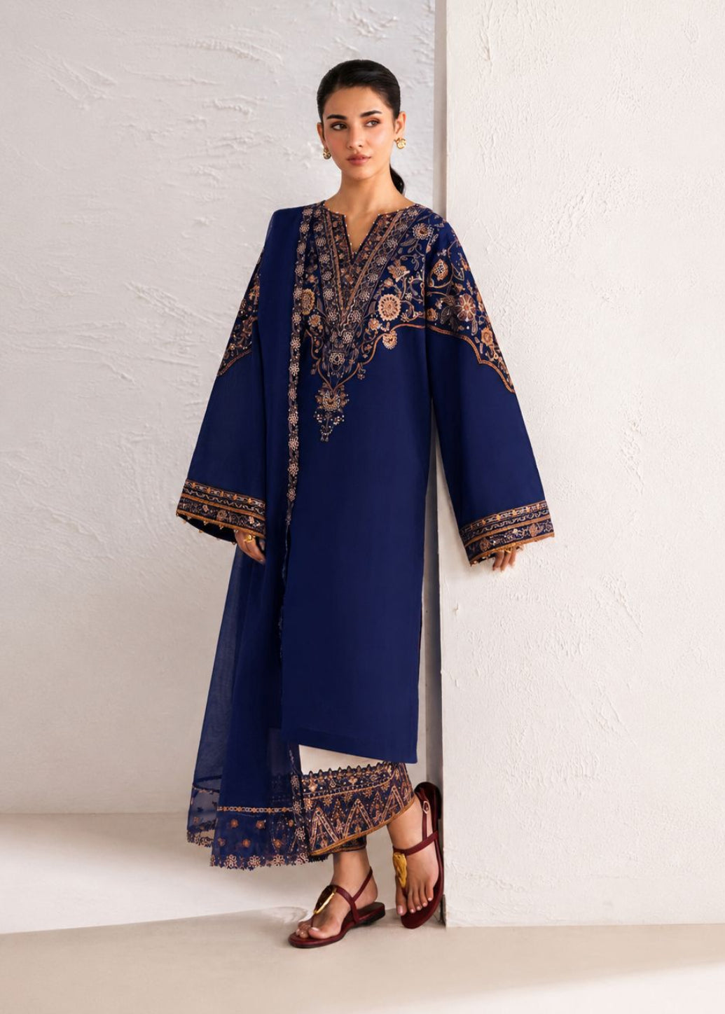 A woman models a navy blue traditional outfit with ornate gold embroidery, standing against a textured off-white wall.