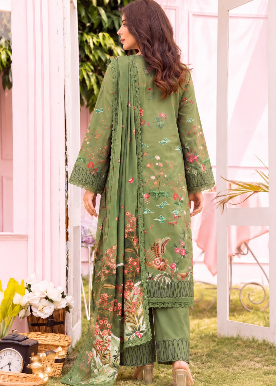 A woman stands outdoors wearing a green embroidered traditional outfit with a long kurta, matching pants, and a dupatta. The fabric features floral and bird motifs in pink, orange, and blue, with green lace trim.