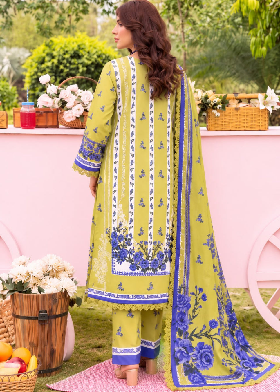A woman wearing a traditional three-piece salwar kameez suit in olive green, featuring blue and white floral embroidery, shown from the back in an outdoor garden setting with a pink backdrop.