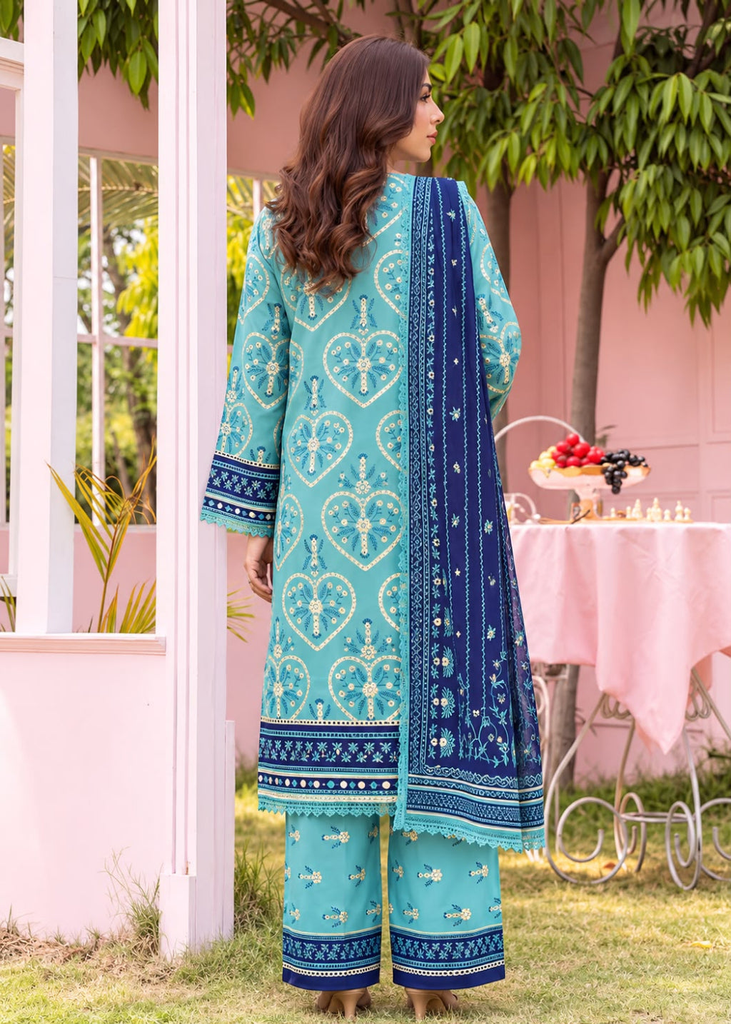 A woman stands outdoors in a garden setting wearing a turquoise and navy printed traditional outfit with matching pants and a draped scarf. The scene includes a pink building, greenery, and a table in the background.
