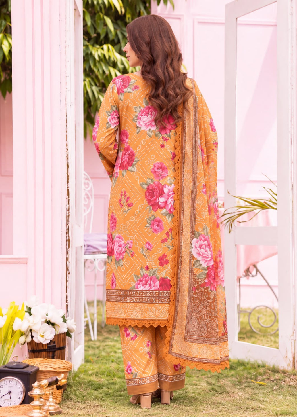 A woman stands outdoors wearing a mustard yellow ethnic outfit with a pink and red floral print, patterned borders, and a matching dupatta. The setting features a pink wall, white door frames, green grass, and decorative props like flowers and a clock.