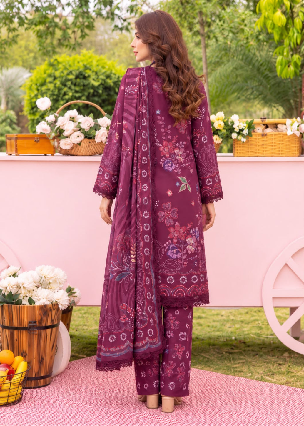 A woman stands outdoors wearing a traditional three-piece salwar kameez suit in deep purple. The outfit features floral embroidery, intricate patterns, and a matching dupatta.