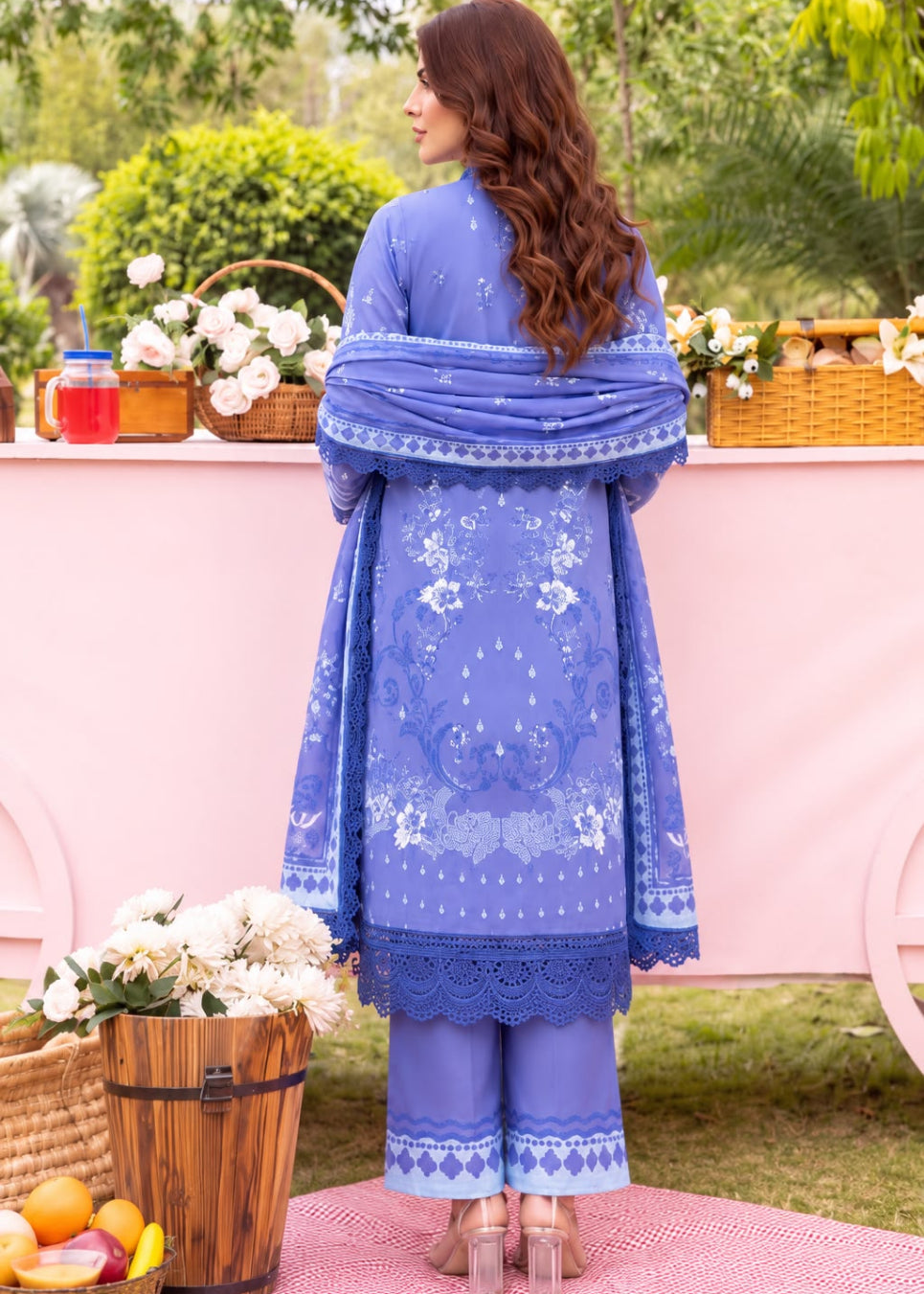 A woman stands outdoors facing away from the camera, wearing a royal blue embroidered traditional outfit with matching dupatta and trousers. The scene includes a pink cart, baskets of flowers, greenery, and fruit in the background.