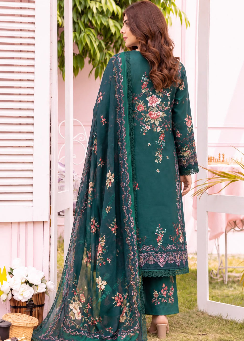 A woman is shown from the back wearing a dark green traditional embroidered outfit with a matching dupatta, set against a soft pink and white outdoor backdrop.