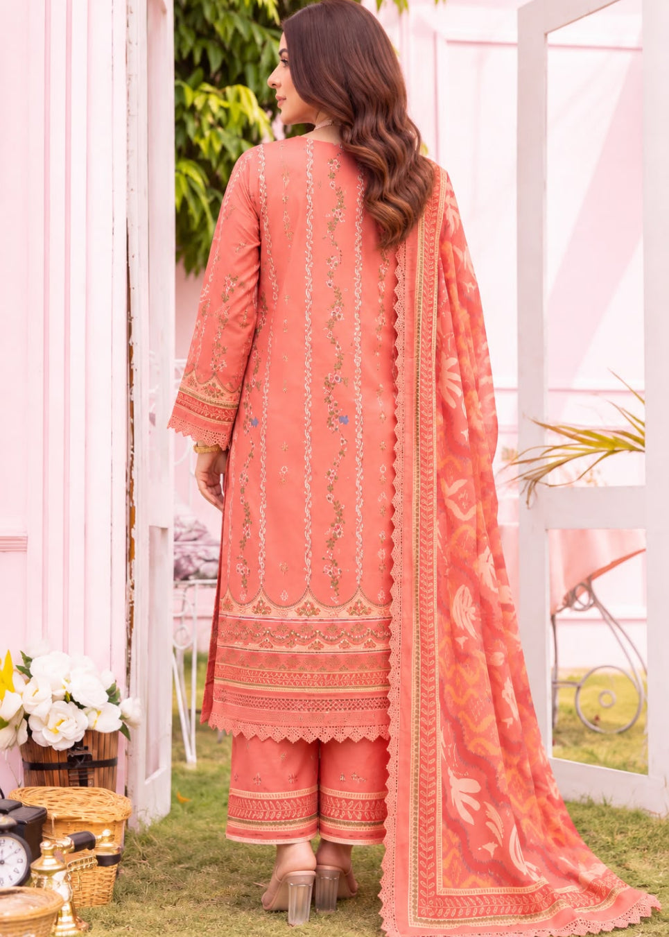 A woman stands outdoors wearing a peach-pink traditional outfit with embroidered details, matching pants, and a long printed dupatta, photographed from the back.