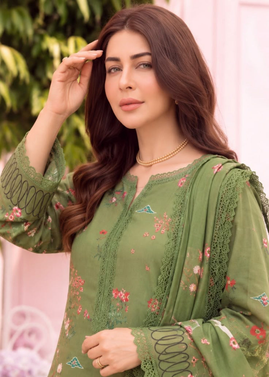 A woman wearing an olive green embroidered ethnic suit with floral patterns and lace details, posed against a soft pink and green background.