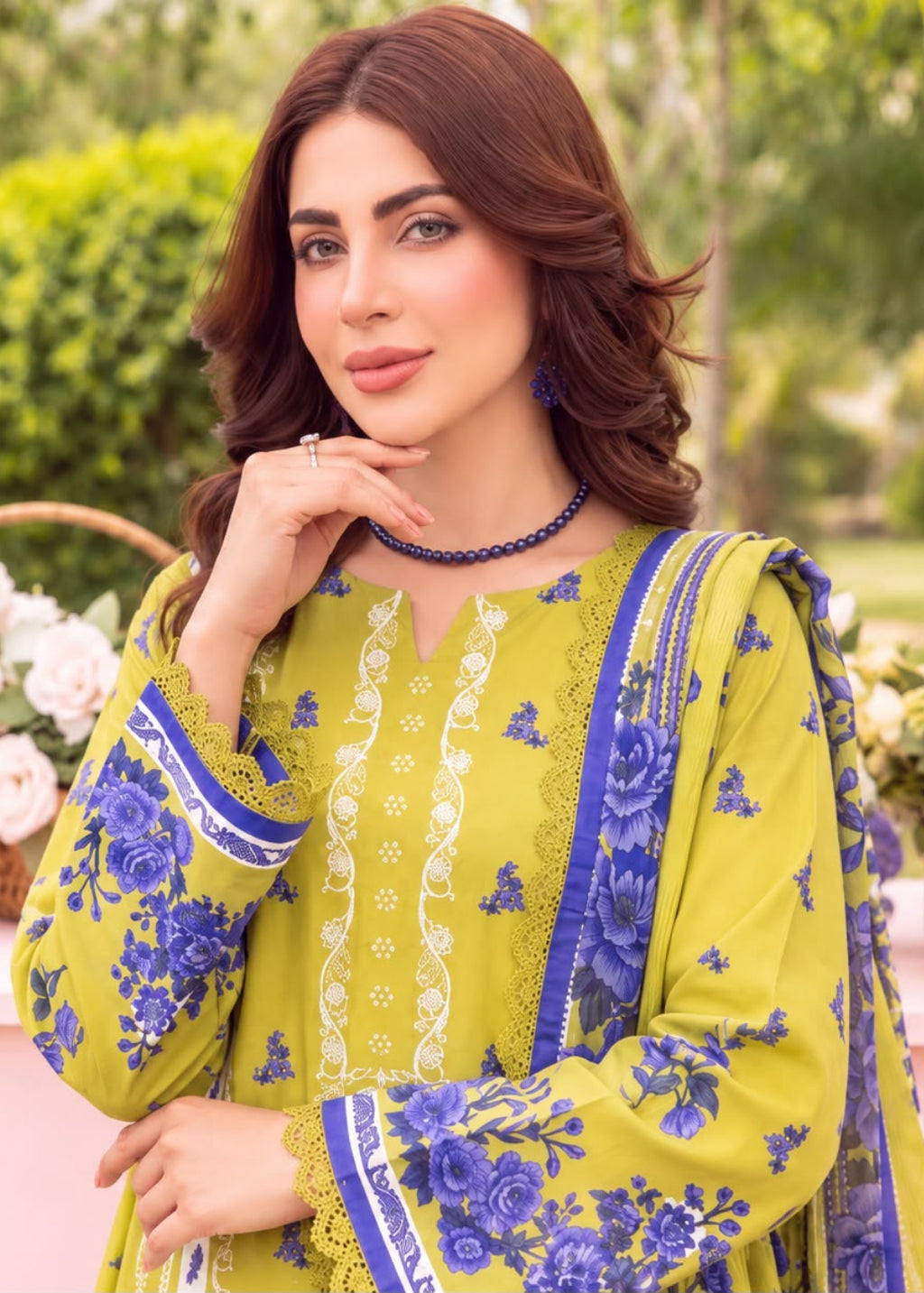A woman wearing a yellow-green traditional embroidered outfit with blue floral patterns and a matching dupatta, posed outdoors in front of a pink table and green garden background.