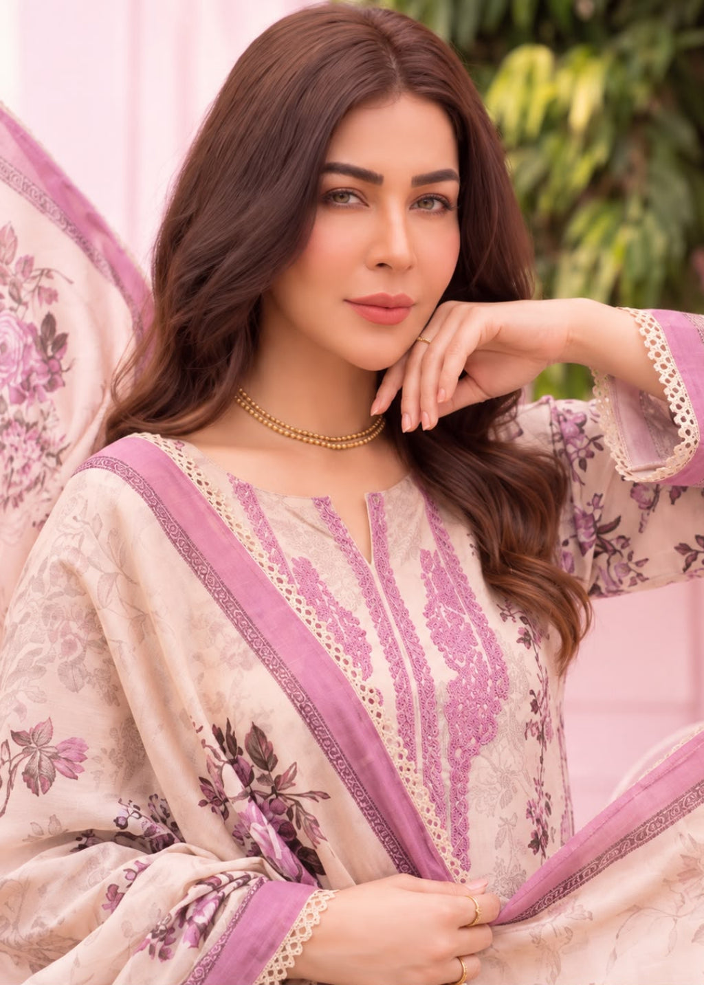 A woman wearing a beige and pink floral printed traditional outfit with embroidered details and a matching dupatta, posed against a soft pink background with green foliage.