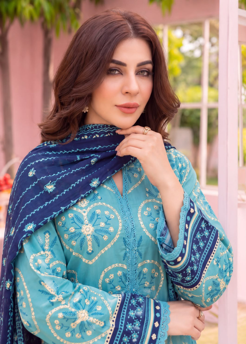 A woman wearing a turquoise embroidered traditional outfit with a navy blue patterned dupatta, posed in front of a soft pink architectural background.